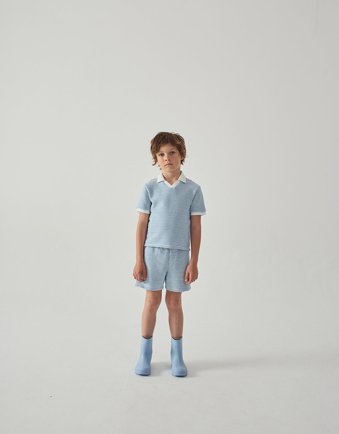 A child stands against a plain white background, wearing a light blue collared shirt, blue socks, and Gingersnaps Boys' Dreyfus Striped Terry Drawstring Shorts.