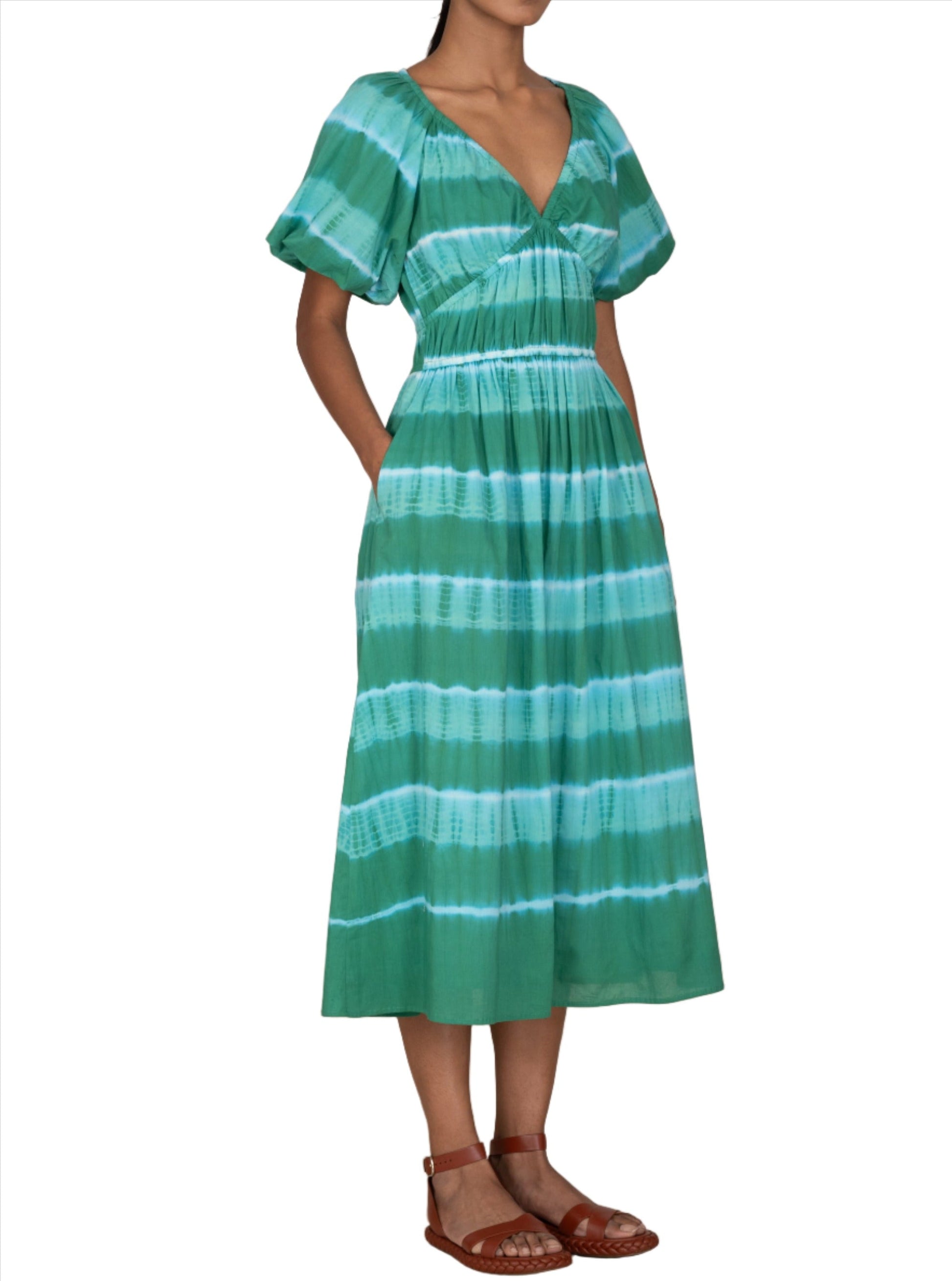 Person wearing an emerald tie-dye Banjanan Francesca Dress with puffed sleeves and a V-neckline. The midi-length dress is paired with brown sandals, and the person’s hands are in the dress pockets.