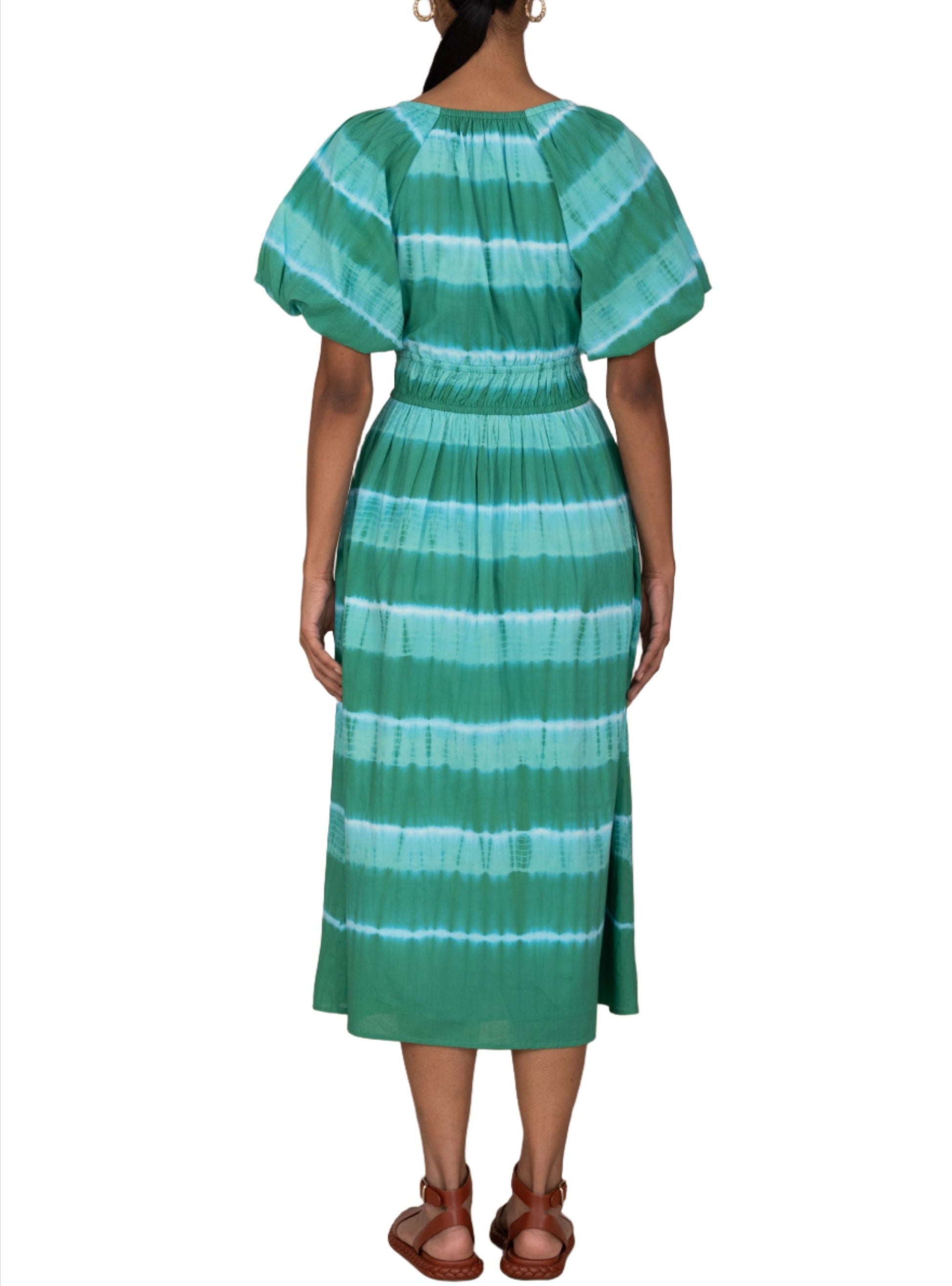 A person wearing a green and blue striped Banjanan Francesca Dress with short sleeves, seen from the back. The midi length dress has a cinched waist and falls below the knees. The person is also wearing brown sandals.