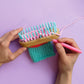 Two hands use the Bright Stripes Nimble Knitter - Squishee Stuffies, an adjustable knitting loom with blue pegs and a hook tool, to knit teal and pink yarn against a purple background.