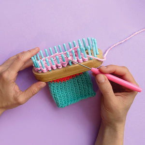Two hands use the Bright Stripes Nimble Knitter - Squishee Stuffies, an adjustable knitting loom with blue pegs and a hook tool, to knit teal and pink yarn against a purple background.