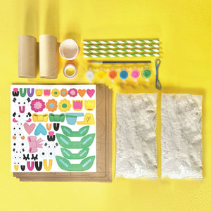 Bright Stripes Paper Mache All Day Kit craft supplies arranged on a yellow surface, featuring cardboard tubes, straws, paints, stickers, string, brown sheets, small cups, tape, and two textured white sheets—perfect essentials for paper mâché projects or creative cardboard sculpting for kids.