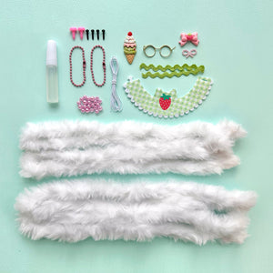 A flat-lay of DIY Chenille Cuties - Bunny Babes craft supplies from Bright Stripes on a light blue background, including glue, beads, doll glasses, a strawberry-print collar, and two lengths of white faux fur trim—perfect components for assembling your own DIY craft kit or making adorable bag danglers.