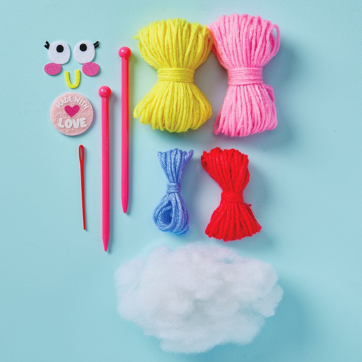 Colorful yarn bundles, plastic needles, a red needle, crafting eyes, a "Made With Love" badge, and polyester stuffing arranged on a light blue background—everything you need to begin knitting projects or start creating with the easy-to-use Snack Knit Kit - Cupcake Cutie from Bright Stripes.