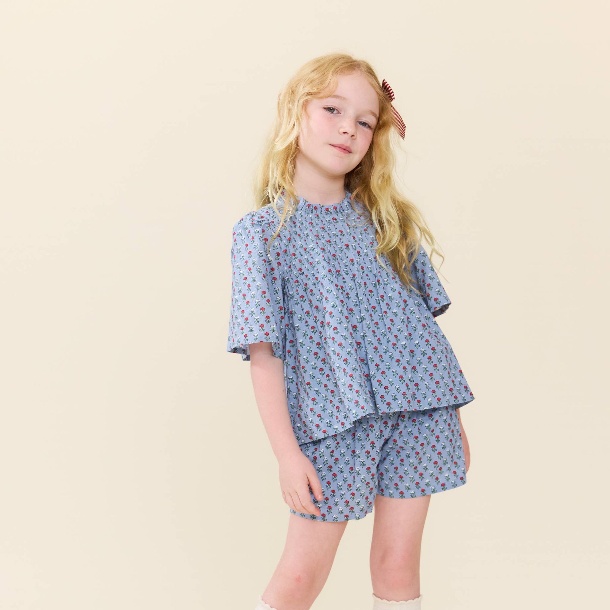 A young girl with long blonde hair is wearing a Pink Chicken blue blockprint patterned top paired with matching Girls’ Theodore Short featuring an elastic waist, standing against a plain light background.