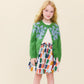 A young girl with long blonde hair wears the Pink Chicken Girls’ Blossom Sweater—a green button-down sweater featuring floral embroidery—paired with a white top and a colorful patterned skirt, standing against a plain light background.