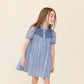 A young girl stands and smiles against a plain light background, wearing the Pink Chicken Girls’ Lucia Dress featuring a blue patterned design, short sleeves, a decorative yoke, and delicate navy smocking.
