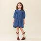 A young girl stands and smiles against a plain, light background, wearing the Girls’ Sawyer Dress by Pink Chicken—a blue chambray style with puff sleeves—paired with a headband and white cowboy boots featuring red detailing.