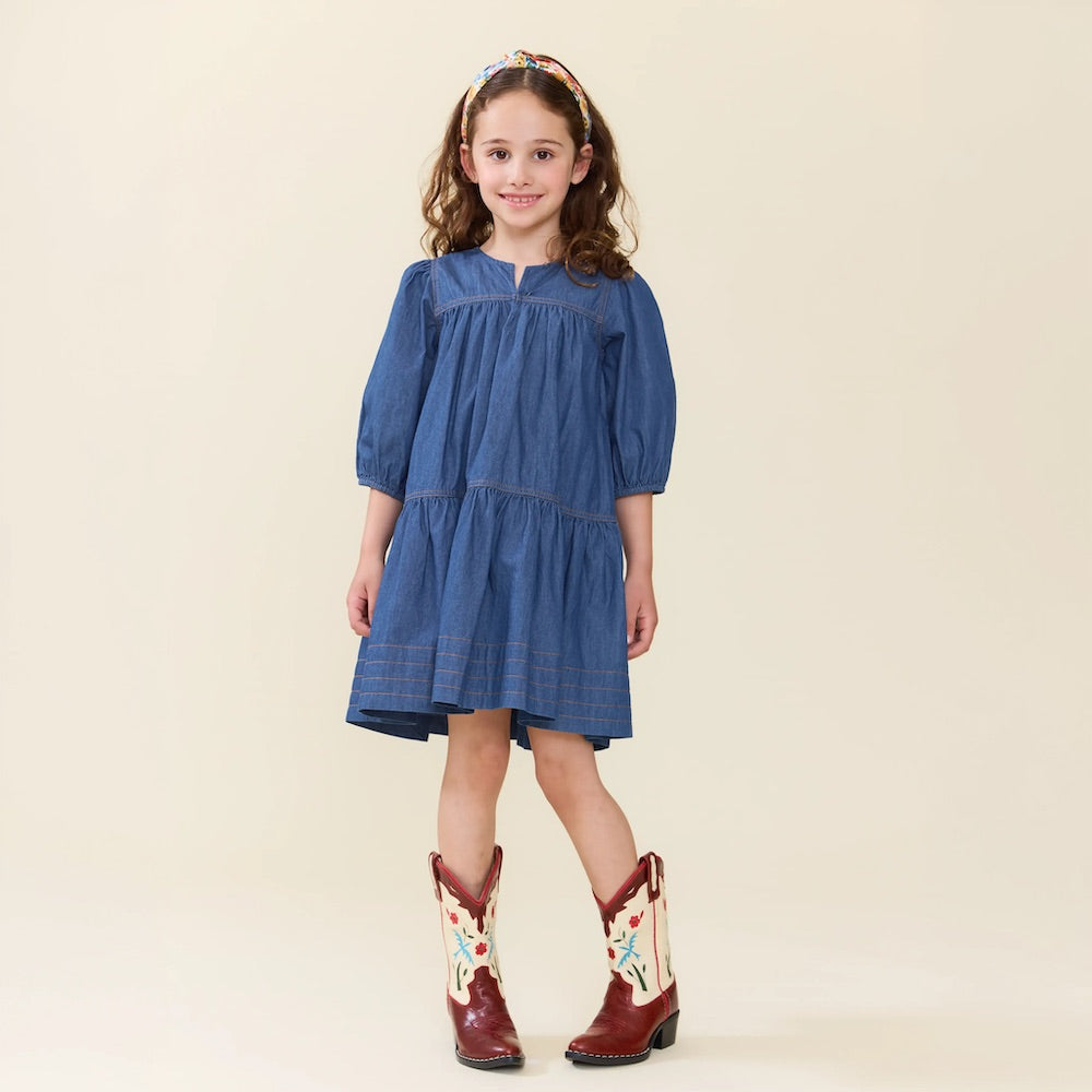 A young girl stands and smiles against a plain, light background, wearing the Girls’ Sawyer Dress by Pink Chicken—a blue chambray style with puff sleeves—paired with a headband and white cowboy boots featuring red detailing.