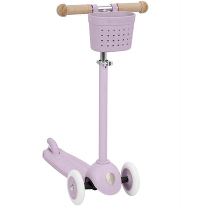 The Banwood Kids’ Eco Scooter with Basket is a lavender three-wheel sustainable scooter for kids, featuring a small front basket, tan handlebar grips, and white wheels, shown on a white background.