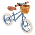 A blue Banwood Kids’ Vintage First Go Balance Bike, featuring white tires, a brown seat and handlebars, and a charming wicker basket on the front—ideal as a child’s first bike for young riders.