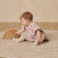 A baby wearing the Quincy Mae Baby Girls' and Girls' Darla Dress with white socks sits on a round rug, playing with wooden rainbow stacking arches on a wooden floor.