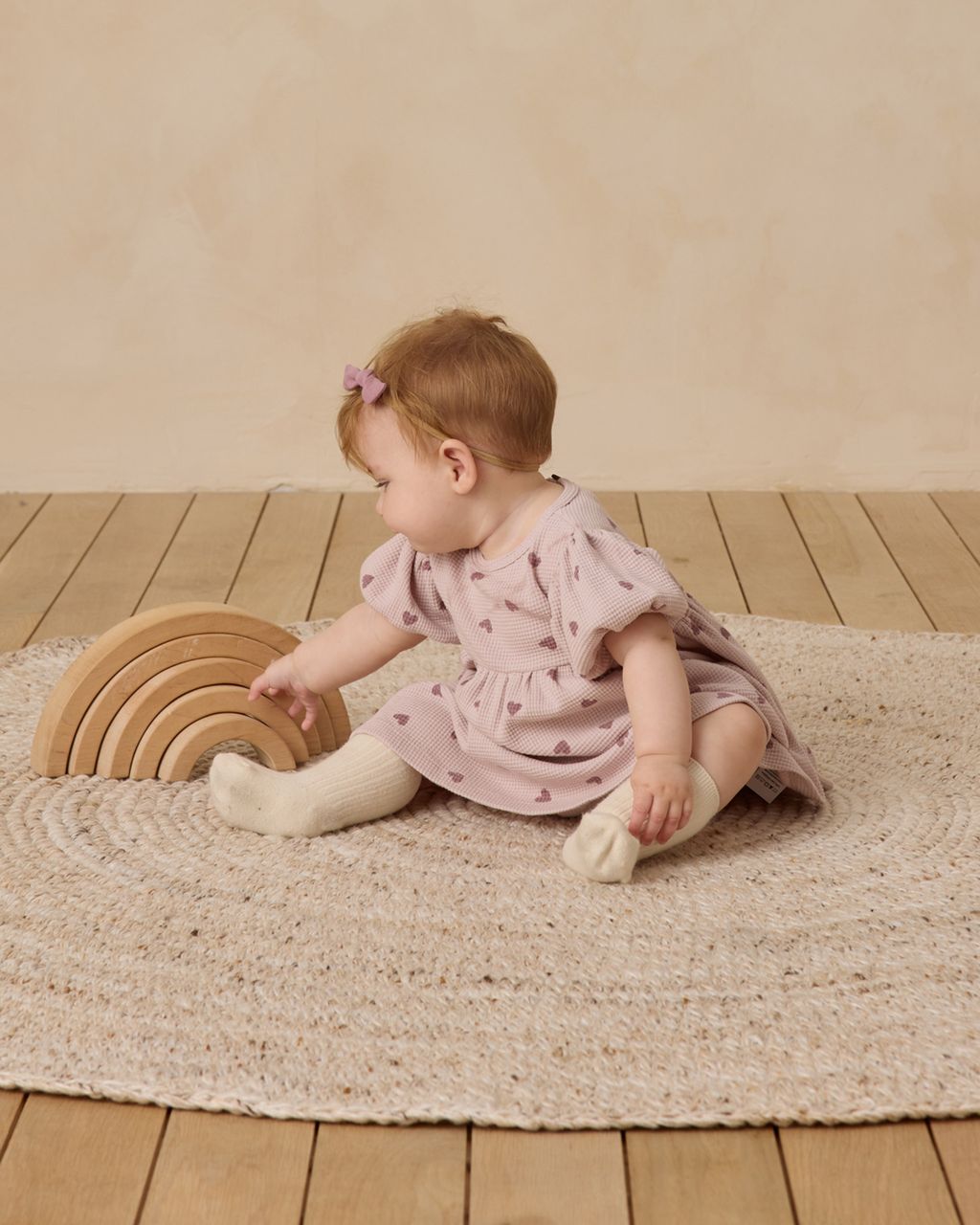 A baby wearing the Quincy Mae Baby Girls' and Girls' Darla Dress with white socks sits on a round rug, playing with wooden rainbow stacking arches on a wooden floor.