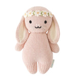 The Baby Bunny - Rose w/ Ivory Floral by Cuddle + Kind is a pink, handcrafted plush bunny made from soft cotton yarn, featuring long ears and an ivory floral crown, displayed against a white background.