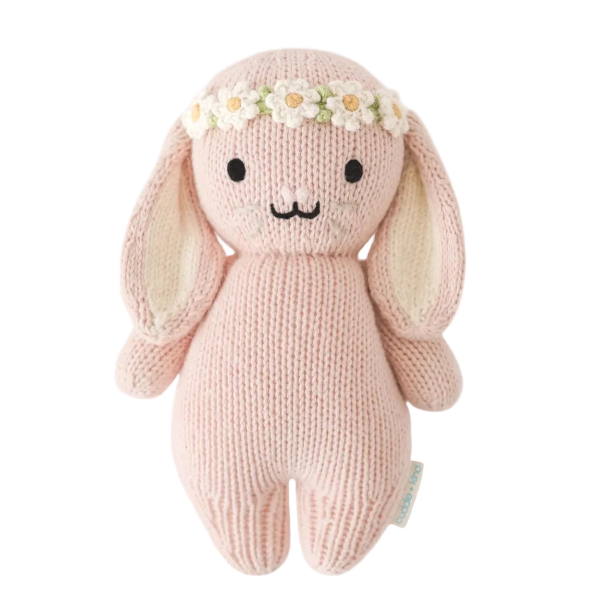 The Baby Bunny - Rose w/ Ivory Floral by Cuddle + Kind is a pink, handcrafted plush bunny made from soft cotton yarn, featuring long ears and an ivory floral crown, displayed against a white background.