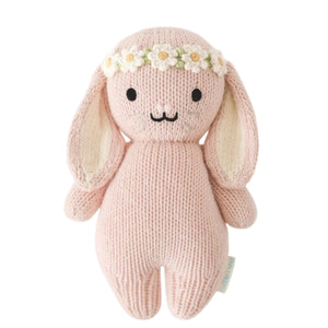 The Baby Bunny - Rose w/ Ivory Floral by Cuddle + Kind is a pink, handcrafted plush bunny made from soft cotton yarn, featuring long ears and an ivory floral crown, displayed against a white background.