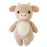 Introducing the Baby Cow by Cuddle + Kind—a beige, handcrafted plush toy made from natural cotton yarn, featuring small horns, round black eyes, and a white belly. This adorable cow is displayed against a white background.