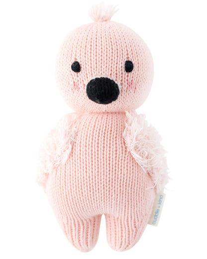 The Baby Flamingo by Cuddle + Kind is a pink hand-knit plush bird with a black beak and eyes, featuring soft textured wings and crafted from Peruvian cotton yarn. It stands upright against a white background.