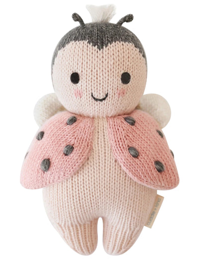 The Baby Ladybug by Cuddle + Kind is an heirloom-quality knitted plush toy featuring a smiling face, pink wings with black spots, and white antennae.