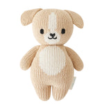Cuddle + Kind’s Baby Puppy, a beige knitted plush dog toy, is handcrafted from cotton yarn and features a white spot on its forehead and belly, charming black eyes, and a small embroidered nose and mouth—this soft baby companion is photographed on a white background.
