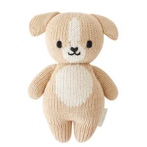 Cuddle + Kind’s Baby Puppy, a beige knitted plush dog toy, is handcrafted from cotton yarn and features a white spot on its forehead and belly, charming black eyes, and a small embroidered nose and mouth—this soft baby companion is photographed on a white background.