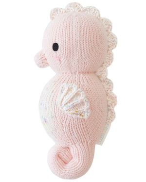 The Baby Seahorse - Powder Pink by Cuddle + Kind is an heirloom-quality plush toy crafted from soft Peruvian cotton yarn, featuring a white fin and tail, black stitched eye, and a small side tag.