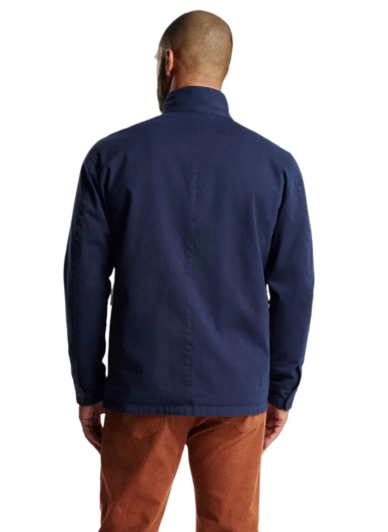 A man stands with his back to the camera, modeling the Peter Millar Stowe Reversible Safari Jacket in navy, paired with brown pants against a stark white background.