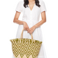 A woman in a white short-sleeve dress holds the Mar Y Sol Bahia Tote, featuring a green and natural zigzag pattern with playful fringe, against a plain white background.