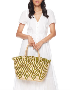 A woman in a white short-sleeve dress holds the Mar Y Sol Bahia Tote, featuring a green and natural zigzag pattern with playful fringe, against a plain white background.