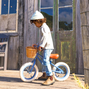 A child wearing a white beanie and boots walks a Banwood Kids’ Vintage First Go Balance Bike with a basket past a weathered wooden building with glass doors.