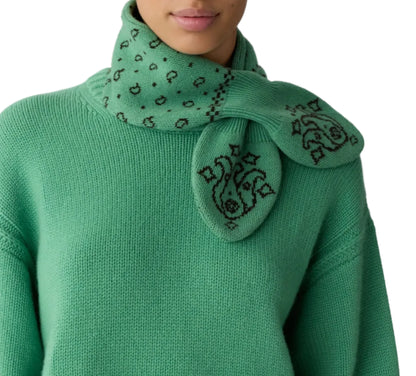 A person is wearing a green knit sweater paired with the Bandana Bowtie Scarf from Guest in Residence, which features black embroidered patterns of abstract faces and shapes. The cozy accessory adds both warmth and style to the look.