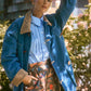 A person stands outdoors, leaning against a tree branch, wearing a blue denim jacket, a blue checkered shirt, and Hunter Bell NYC Ashton Short in Olive Floral linen with a tailored banded waist.