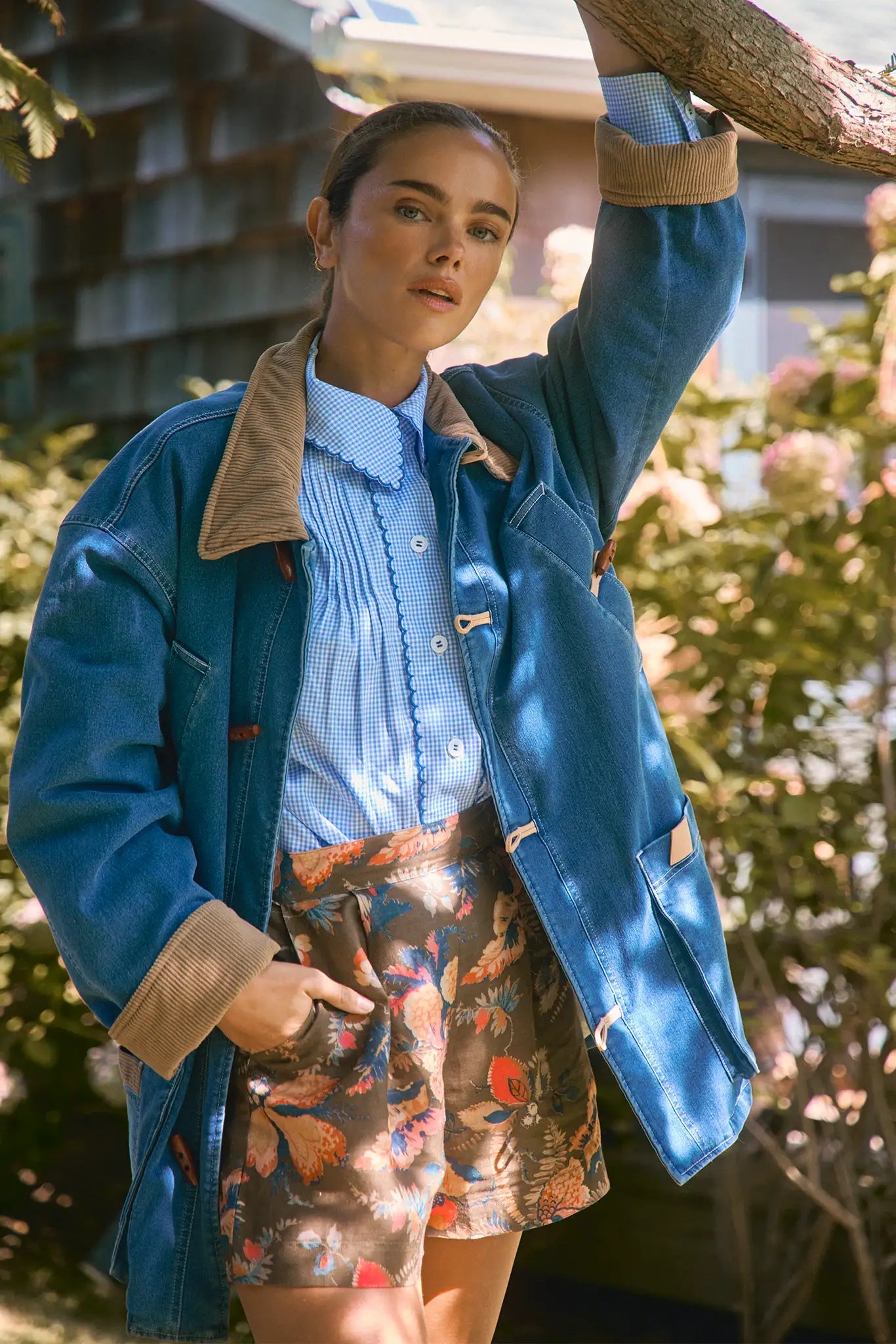 A person stands outdoors, leaning against a tree branch, wearing a blue denim jacket, a blue checkered shirt, and Hunter Bell NYC Ashton Short in Olive Floral linen with a tailored banded waist.