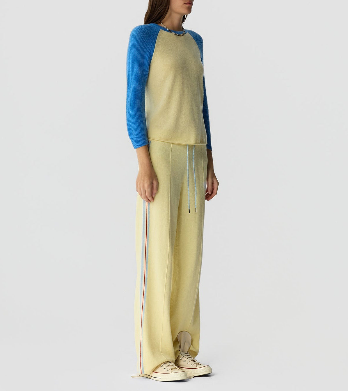 A person wears the Guest in Residence Baseball Tee—a lightweight cashmere beige and blue colorblock raglan top—paired with matching beige wide-leg pants with side stripes and beige sneakers, standing against a plain light background.