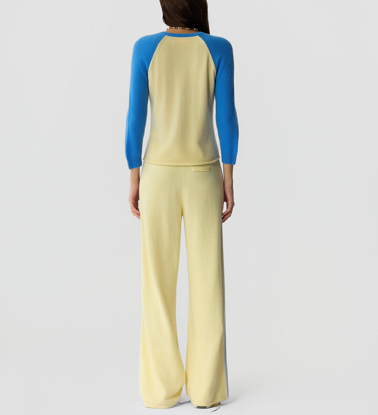 A person stands facing away, wearing Guest in Residence's Baseball Tee with blue colorblock raglan sleeves and wide-leg yellow pants.