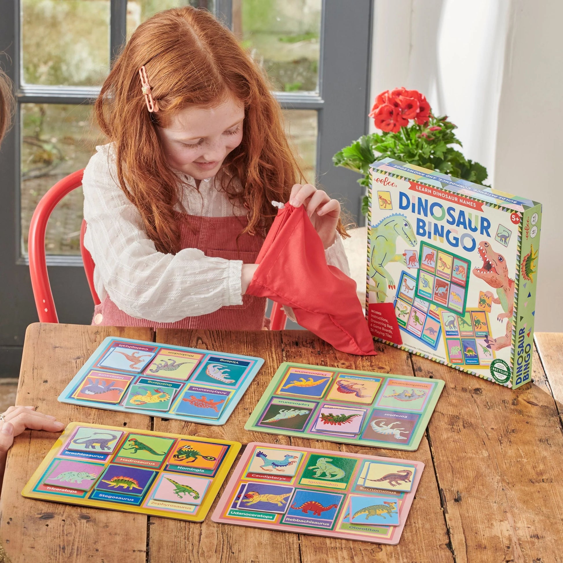 A child plays Eeboo Dinosaur Bingo at a table, drawing pieces from a red bag while several bingo cards and the game box are visible.