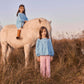 Two young girls wearing Louise Misha’s Girls’ Risa Blouse pose with a white horse in a grassy field; one sits atop the horse while the other stands in front. Their cotton and linen blouses, adorned with delicate floral embroidery and charming puffy sleeves, add a whimsical touch to the tranquil scene.