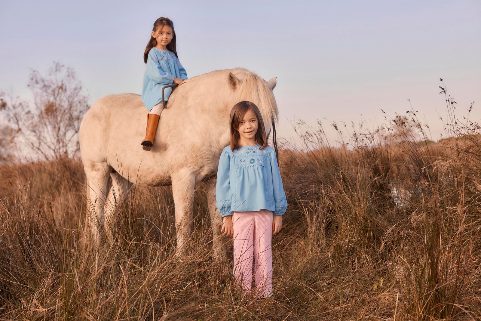 Two young girls wearing Louise Misha’s Girls’ Risa Blouse pose with a white horse in a grassy field; one sits atop the horse while the other stands in front. Their cotton and linen blouses, adorned with delicate floral embroidery and charming puffy sleeves, add a whimsical touch to the tranquil scene.