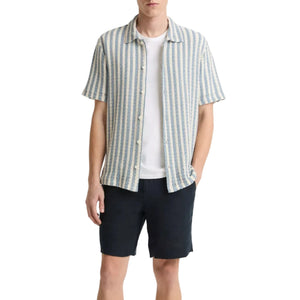A man wearing the vince Playa Stripe Shortsleeve Shirt over a white tee with dark shorts, standing with one hand in his pocket against a white background.