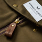 A close-up view of a brown jacket showcasing a gold zipper with a leather pull tab and featuring a white label that reads "Bennett Winch," recalling their famous S.C Holdall collection.