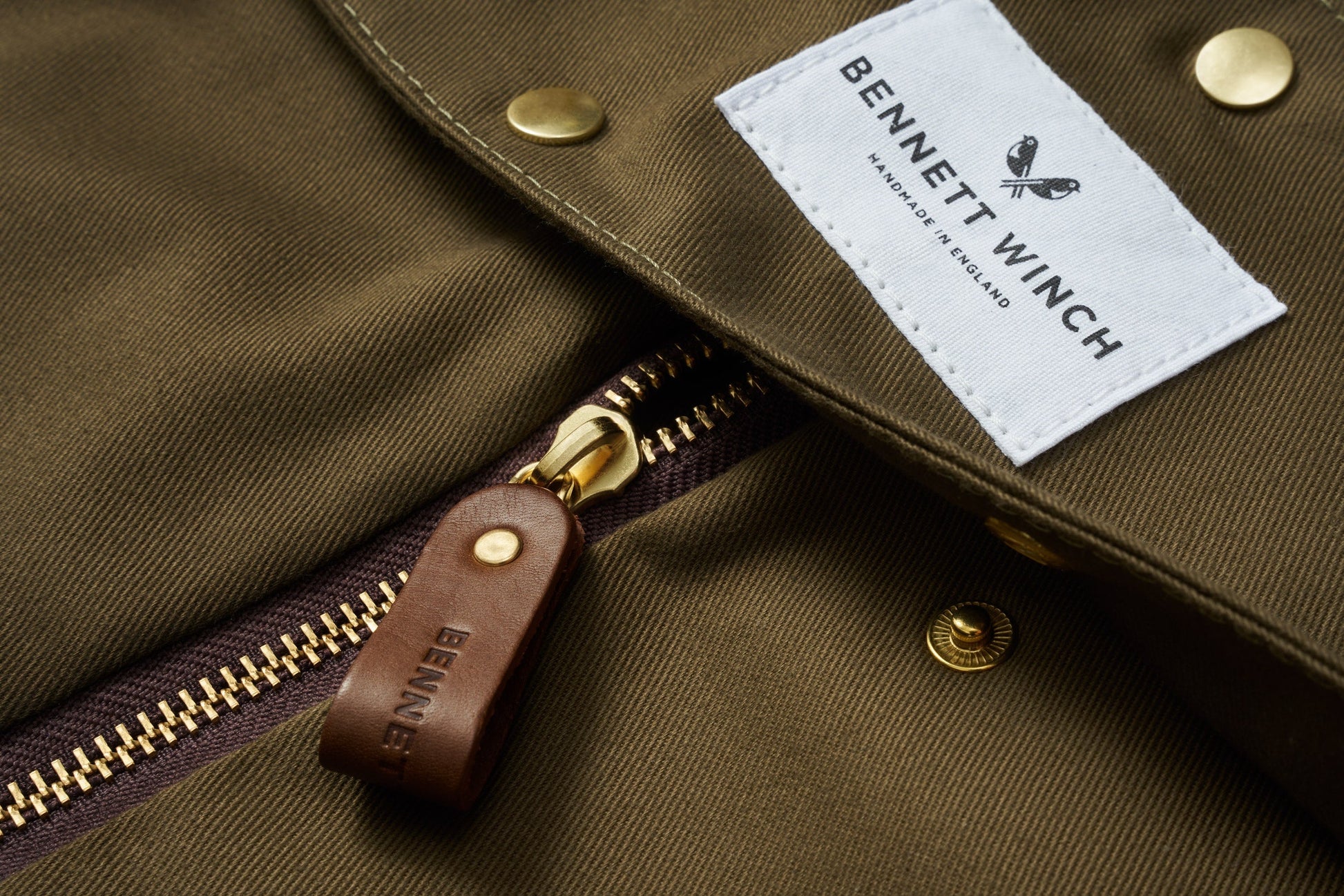 A close-up view of a brown jacket showcasing a gold zipper with a leather pull tab and featuring a white label that reads "Bennett Winch," recalling their famous S.C Holdall collection.