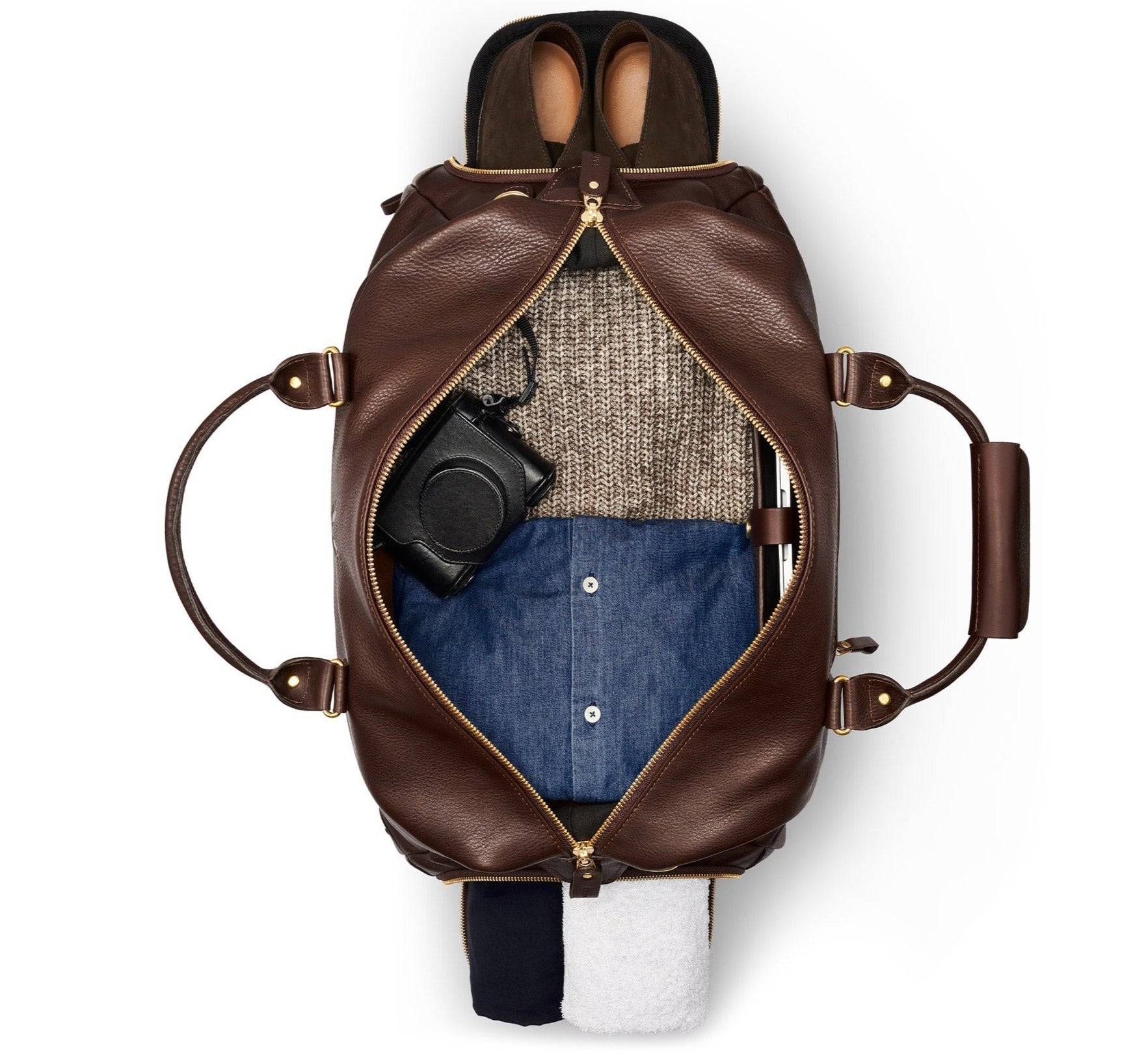 A top view of an open Bennett Winch Leather Weekender reveals a gray sweater, blue shirt, black pants, white towel, black camera, and brown boots inside. This premium travel bag from Bennett Winch is crafted from full-grain leather, offering a perfect blend of style and functionality for the discerning traveler.