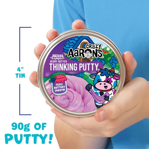 A person holds a 4-inch tin of Crazy Aaron’s Better Butter - Berry Thinking Putty, a berry-scented putty perfect for sensory play. The label features a cartoon cow and text indicating it contains 90g of Thinking Putty.