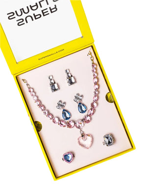 The Super Smalls Big Presentation Jewelry Set features a yellow box with a pink beaded heart necklace, three earring pairs, and two adjustable rings decorated with pink, blue, and silver stones for pastel elegance.