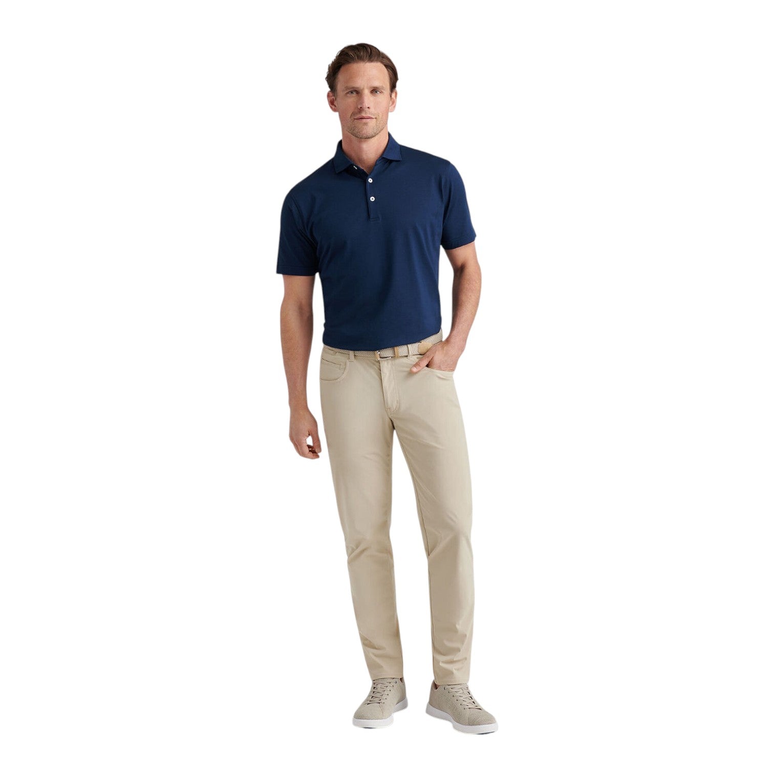 A man faces forward wearing the Peter Millar Bingham Performance Five-Pocket Pant in beige, a navy blue wrinkle-resistant Italian knit polo shirt, and light gray sneakers against a white background.