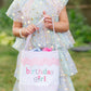 A young girl in a pastel, star-patterned dress holds a Bits & Bows Birthday Girl Tote labeled "birthday girl" outdoors.