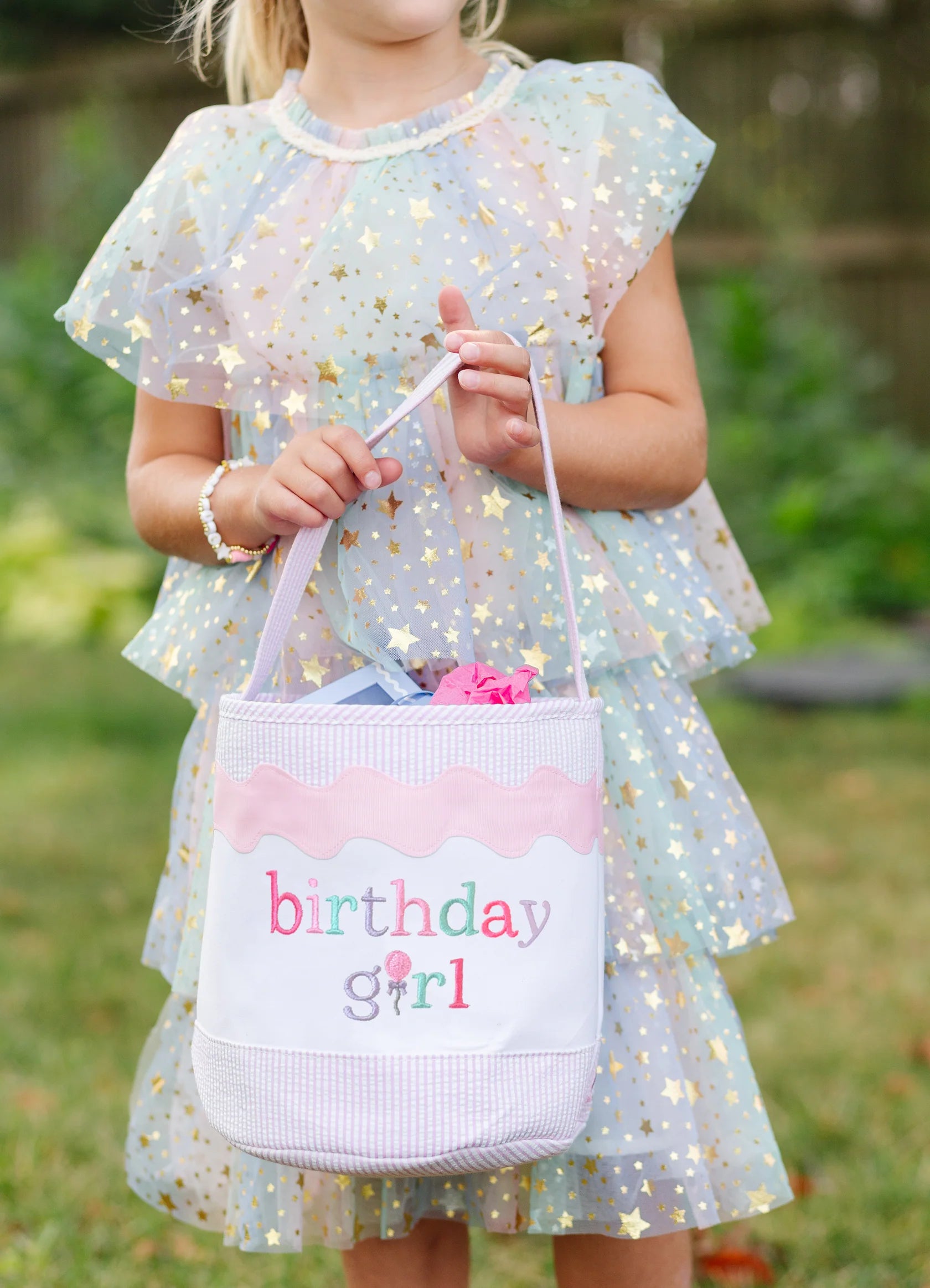 A young girl in a pastel, star-patterned dress holds a Bits & Bows Birthday Girl Tote labeled "birthday girl" outdoors.