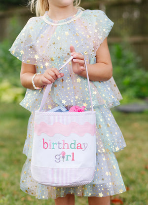 A young girl in a pastel, star-patterned dress holds a Bits & Bows Birthday Girl Tote labeled "birthday girl" outdoors.