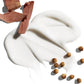 A smear of white cream, identified as Jack Black Black Reserve Hydrating Body Lotion and enriched with nourishing Shea Butter, is displayed on a white background, accompanied by several brown seeds and pieces of light brown wood.