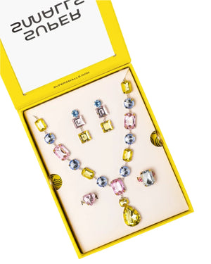 The Super Smalls Black Tie Mega Set features a necklace, earrings, and an adjustable ring with oversized pink, yellow, and blue gems, all displayed in an open yellow box.